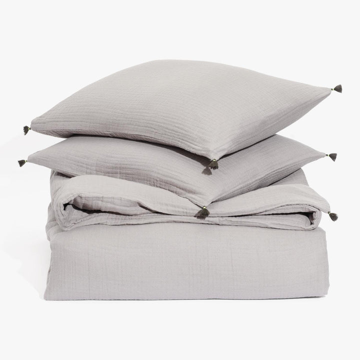 Grey textile bedding set with pillows and tassels, neatly stacked