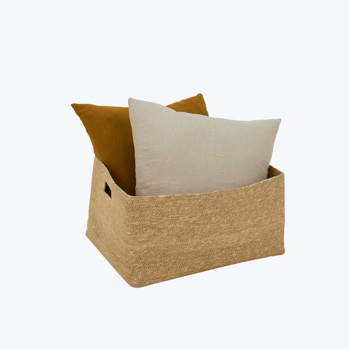 Rectangular jute basket with cut-out handles holding two pillows.