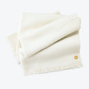 Neatly folded cream-colored textile with fringe detailing and small emblem.