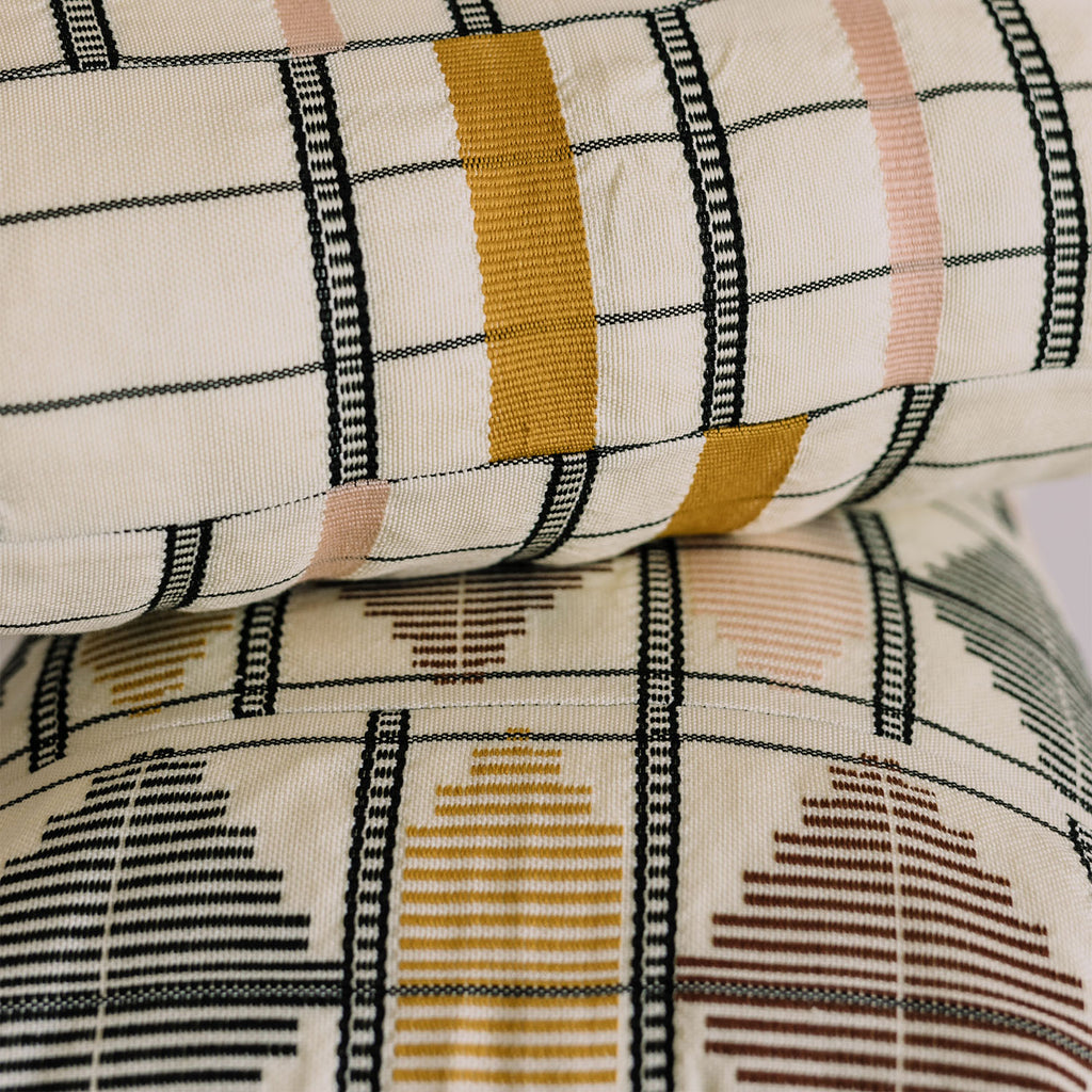 Close-up of decorative pillows with textured fabric patterns in black, mustard yellow, pale pink, brown, yellow, and black against off-white background.