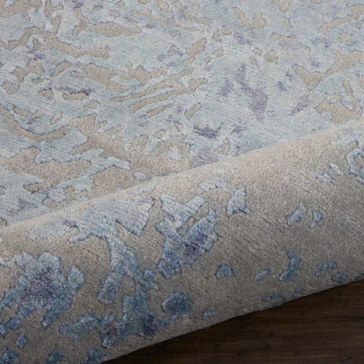 Close-up of plush area rug with rolled edge in muted grays and blues, featuring an abstract, nature-inspired pattern; rests on smooth wooden floor.